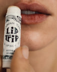 A close-up of a person holding an uncapped tube of Faire Skin Trip Lip Balm near their parted lips, ready to apply the moisturizing balm.