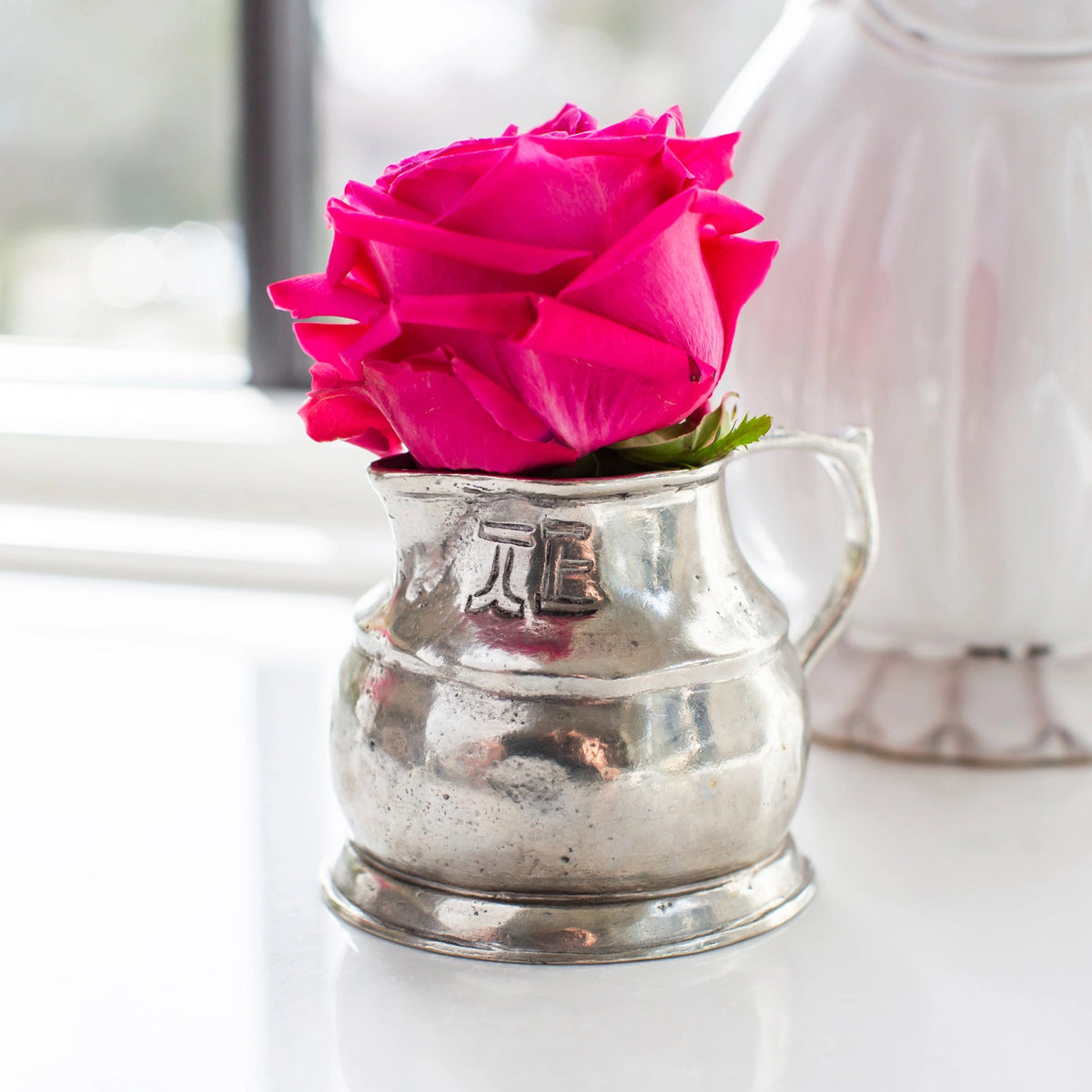 A vibrant pink rose rests in Faire’s Vintage 1815 Pitcher, a small, handmade piece with engraved Italian detailing, gleaming softly on a white surface by the window.