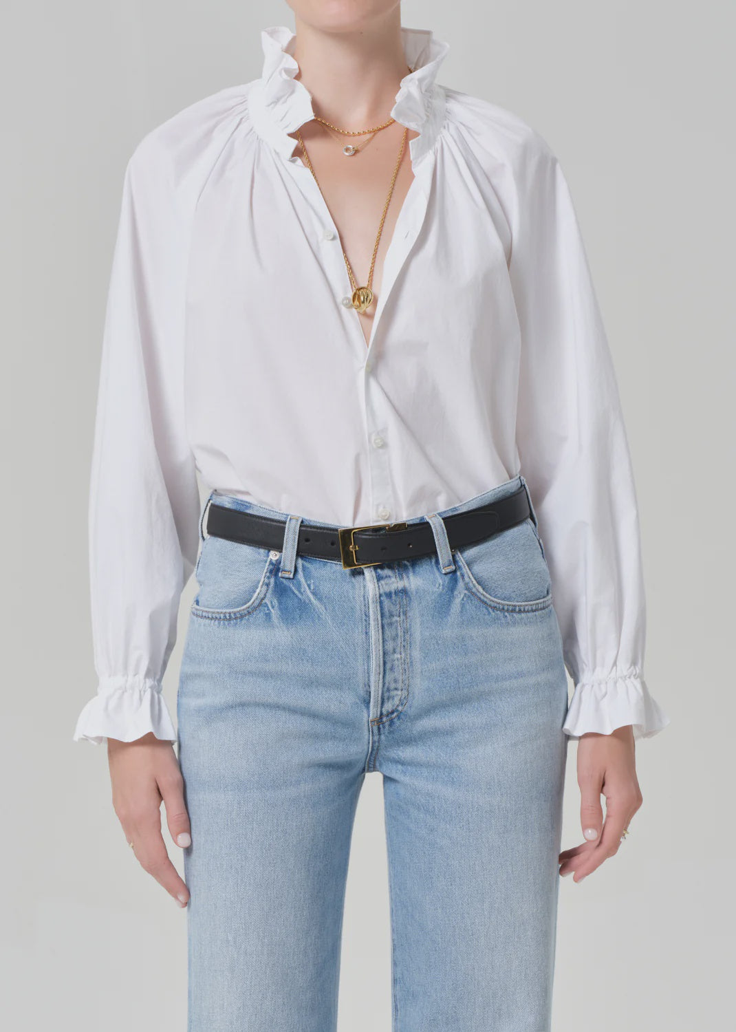 A person wears the loretta ruffle top in optic white by Citizens Of Humanity/AGOLDE, styled with light blue high-waisted jeans, a black belt, layered gold necklaces, and ruffled cuffs, standing against a plain light background.
