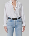A person wears the loretta ruffle top in optic white by Citizens Of Humanity/AGOLDE, styled with light blue high-waisted jeans, a black belt, layered gold necklaces, and ruffled cuffs, standing against a plain light background.