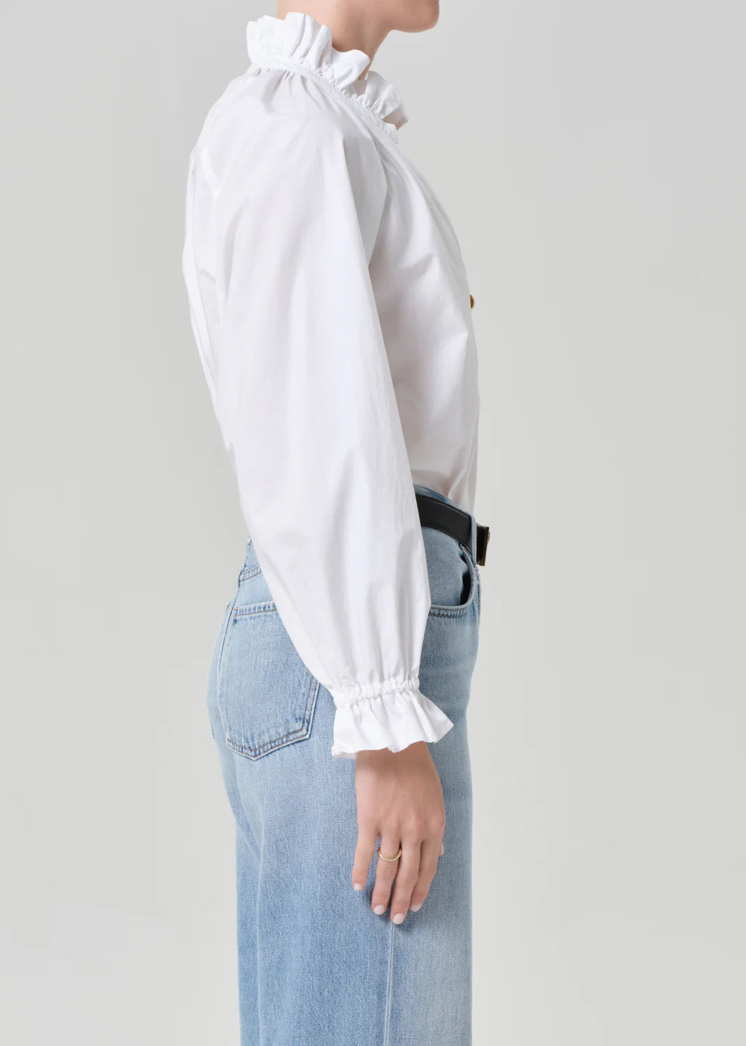 A person stands in profile wearing the Citizens Of Humanity/AGOLDE loretta ruffle top in optic white, styled with light blue high-waisted jeans against a plain light gray background.