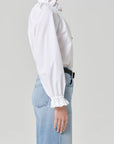 A person stands in profile wearing the Citizens Of Humanity/AGOLDE loretta ruffle top in optic white, styled with light blue high-waisted jeans against a plain light gray background.