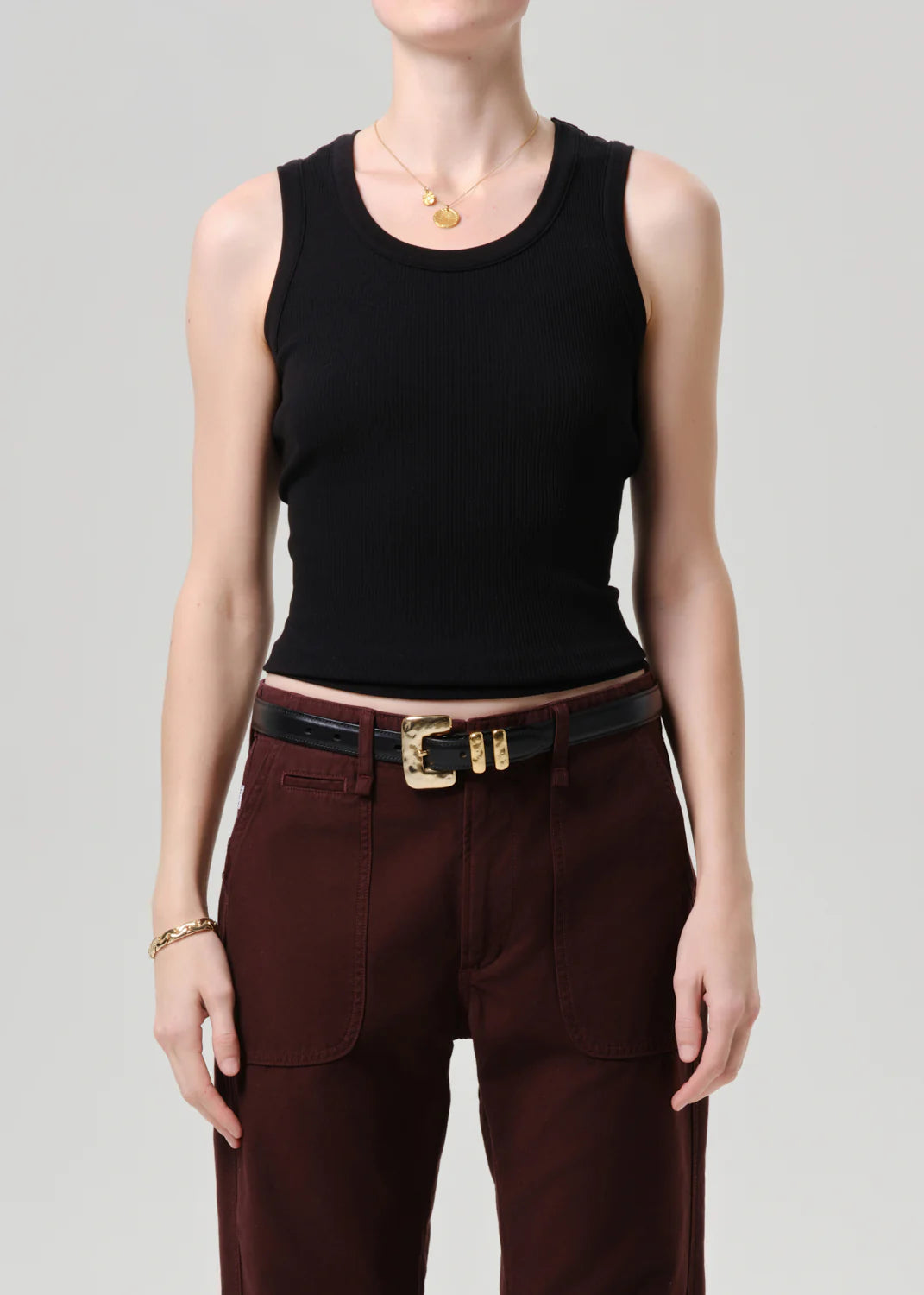 A person wearing the Citizens Of Humanity/AGOLDE nova tank, styled with high-waisted dark red pants, a black belt with a gold buckle, and gold jewelry, stands against a plain light background.