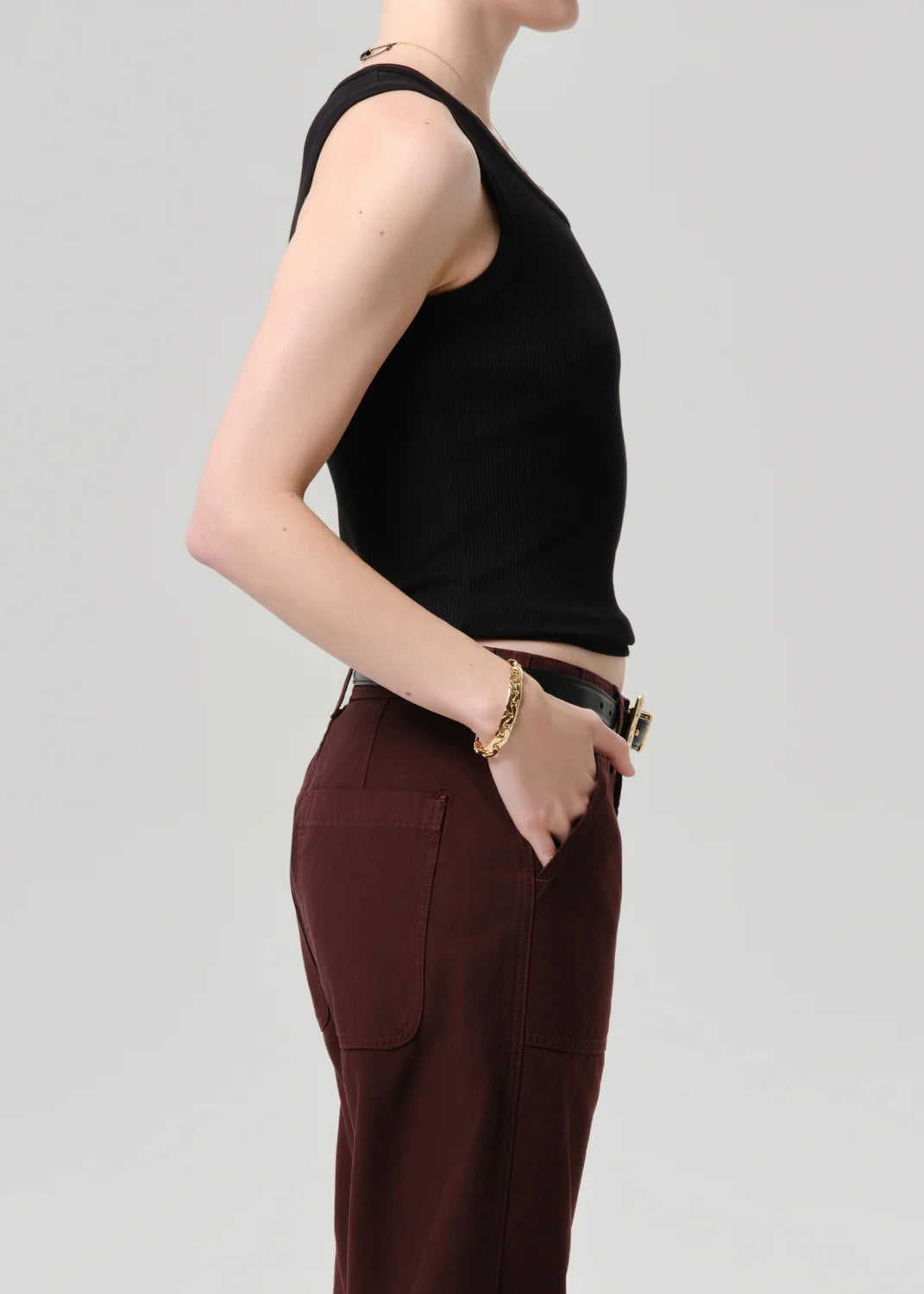 A person stands in profile against a plain background, wearing the Citizens Of Humanity/AGOLDE nova tank with a scoop neckline and high-waisted maroon pants. One hand is in their pocket, accessorized with a bracelet and gold-buckle belt.