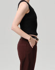 A person stands in profile against a plain background, wearing the Citizens Of Humanity/AGOLDE nova tank with a scoop neckline and high-waisted maroon pants. One hand is in their pocket, accessorized with a bracelet and gold-buckle belt.