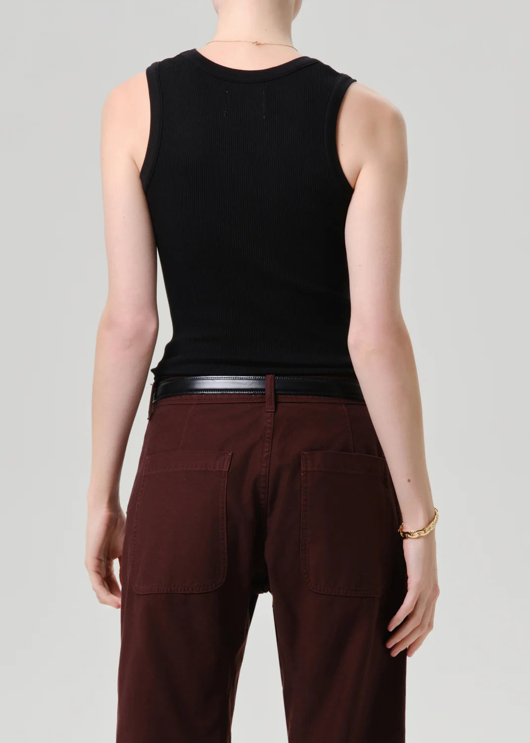 A person is shown from the back wearing the Citizens Of Humanity/AGOLDE nova tank in black with dark brown pants, a black belt, a gold bracelet, and a thin necklace against a plain light gray background.