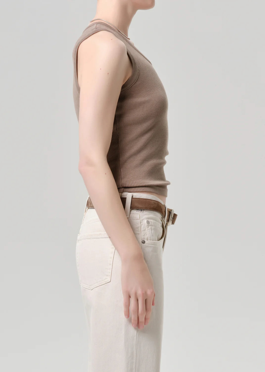 A person stands in profile against a plain background, wearing the Citizens Of Humanity/AGOLDE nova tank—a taupe ribbed top with a scoop neckline—paired with light pants and a brown belt. Their face is not visible.