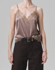 A model wears the Citizens Of Humanity/AGOLDE lace trimmed cami in mauve Japanese hammered satin, tucked into high-waisted black pants with a belt, accessorized with a short necklace and bracelet against a plain light gray background.