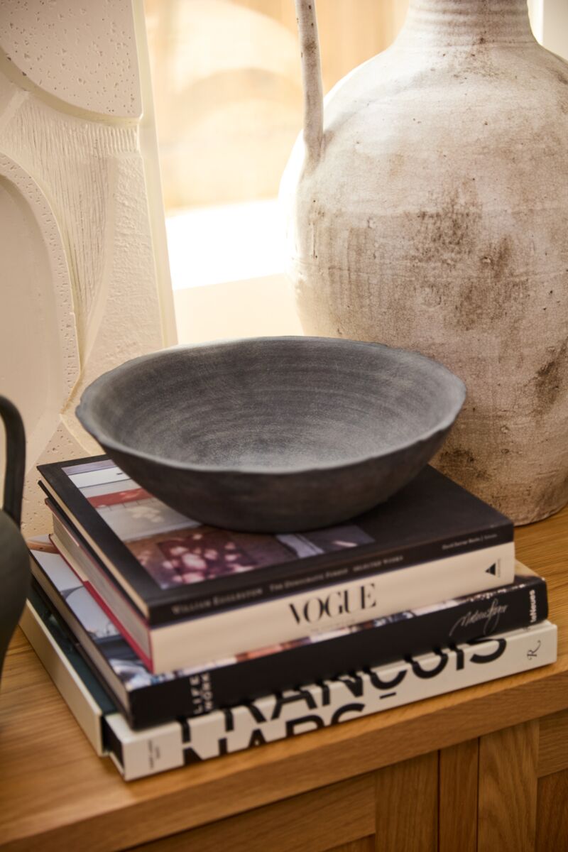 The Rossetti Bowl SM by Accent Decor in matte black rests on a stack of art and fashion books, including Vogue, beside a large textured white vase on a wooden surface in a bright, stylish interior.