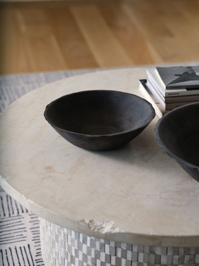 The Rossetti Bowl SM by Accent Decor in matte black sits on a round, light stone table beside books, with another handcrafted terracotta bowl and a patterned rug visible in the background.