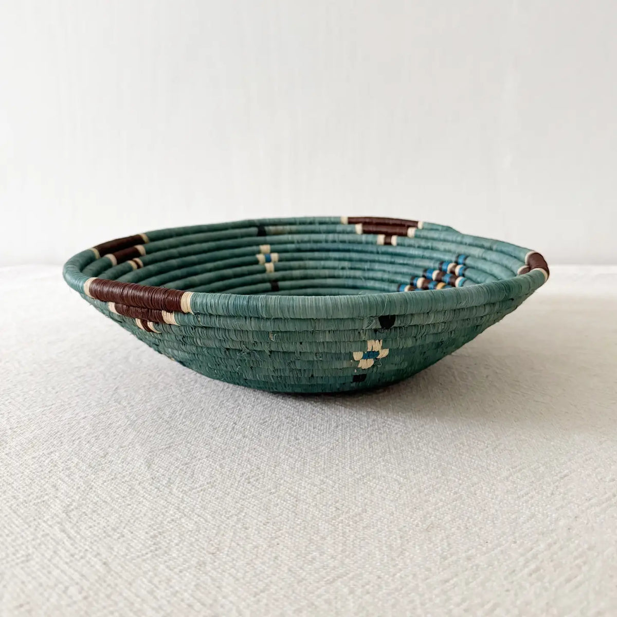 The Birava Large Bowl by Faire is a shallow, round woven basket in teal, brown, cream, and blue—ideal as handmade decor or Rwanda wall art. Shown on a light textured surface with a plain white background.
