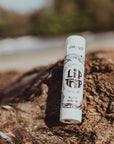 A tube of Faire Skin Trip Lip Balm stands upright on textured driftwood against a blurred natural background. The label says "All Day Moisture," highlighting the soothing, natural benefits of this lip balm.