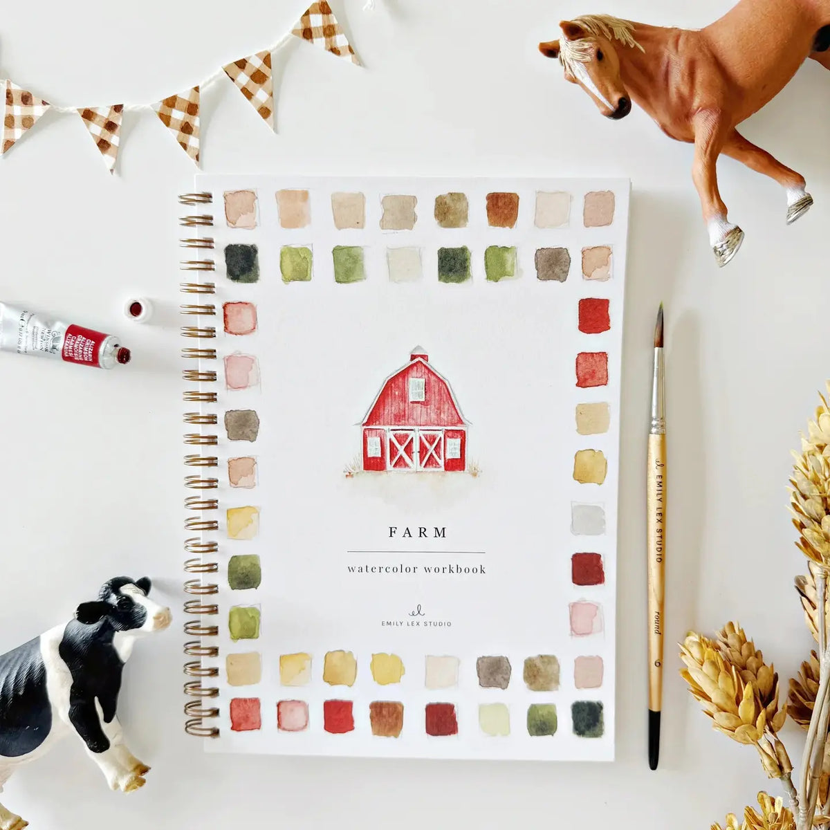 The Faire Watercolor Workbook is a beginner-friendly, spiral-bound book with a red barn cover featuring paint swatches. Nearby are a paintbrush, tube of paint, toy horse, cow, wheat stalks, and checkered bunting.