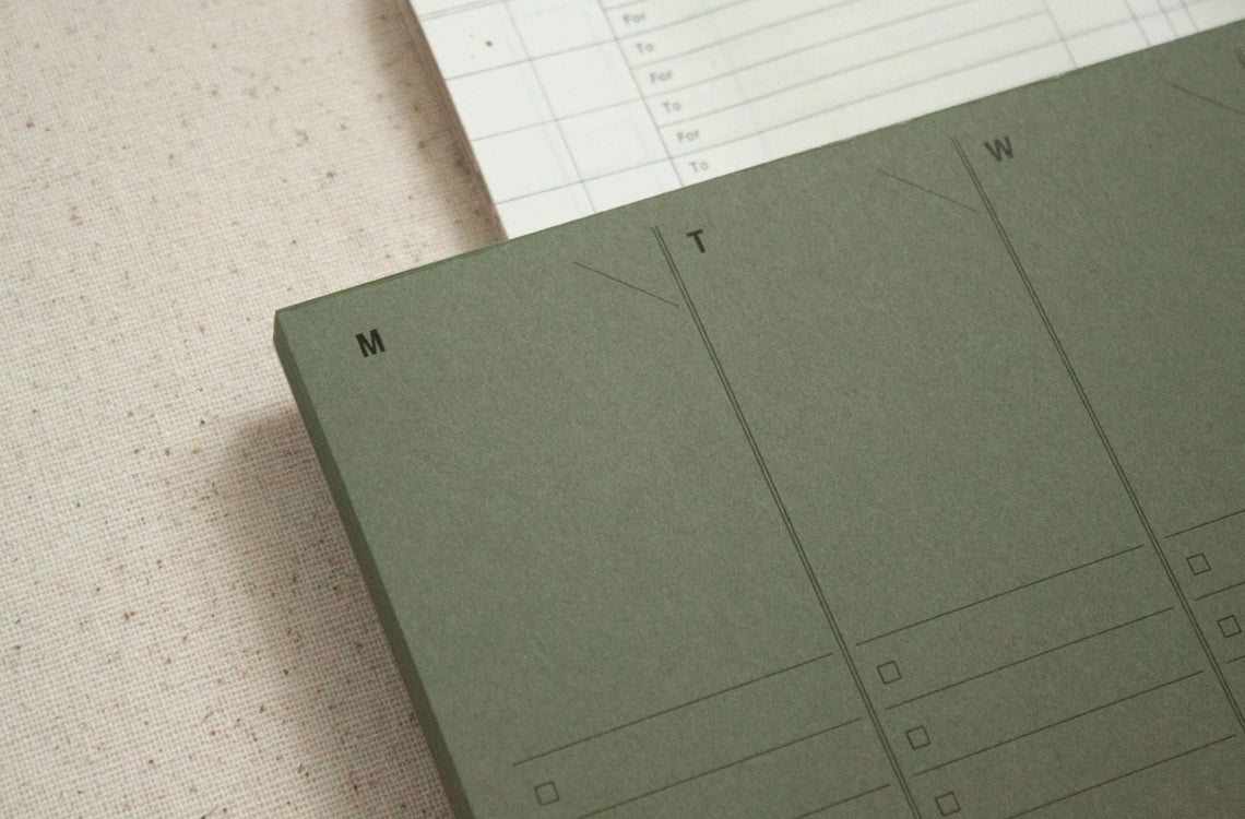 A close-up of the Faire Weekly Planner Paper Notepad in green displays "M" and "T" columns for Monday and Tuesday with checklist boxes. A second lined sheet appears partially above the planner.