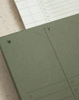 A close-up of the Faire Weekly Planner Paper Notepad in green displays "M" and "T" columns for Monday and Tuesday with checklist boxes. A second lined sheet appears partially above the planner.