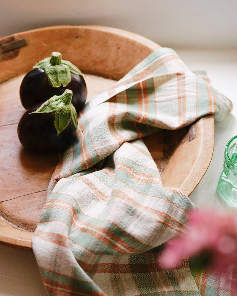 Two small eggplants sit on a wooden tray beside the Faire Cook's Cloth, a pastel plaid cotton-linen kitchen towel. A green glass and a blurred pink flower appear at the edges of the image.