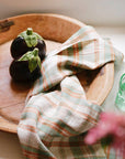 Two small eggplants sit on a wooden tray beside the Faire Cook's Cloth, a pastel plaid cotton-linen kitchen towel. A green glass and a blurred pink flower appear at the edges of the image.