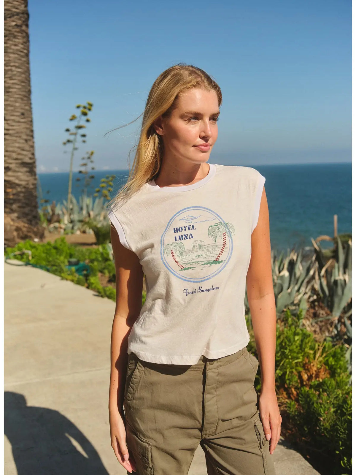 A woman with blonde hair wears the Hotel Luna Muscle Tank by Faire and khaki pants as she walks outdoors along a coastline, with blue ocean, sky, and green plants in the background.