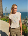 A woman with blonde hair wears the Hotel Luna Muscle Tank by Faire and khaki pants as she walks outdoors along a coastline, with blue ocean, sky, and green plants in the background.