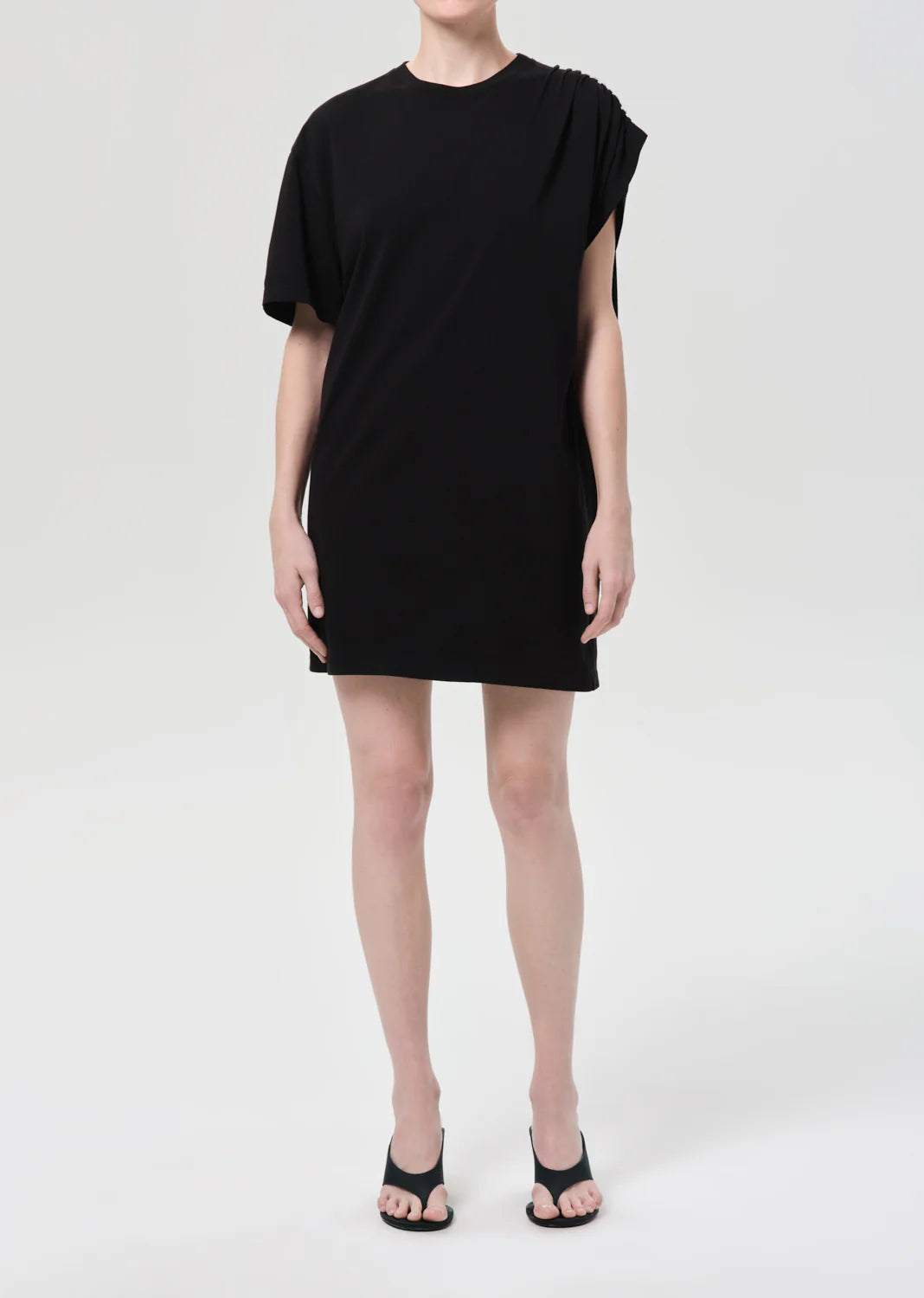 A person stands against a plain background wearing the Citizens Of Humanity/AGOLDE Roma Dress, a loose-fitting black organic cotton mini dress with an asymmetrical drape on one shoulder and short sleeves, paired with black open-toe heels. Face not visible.