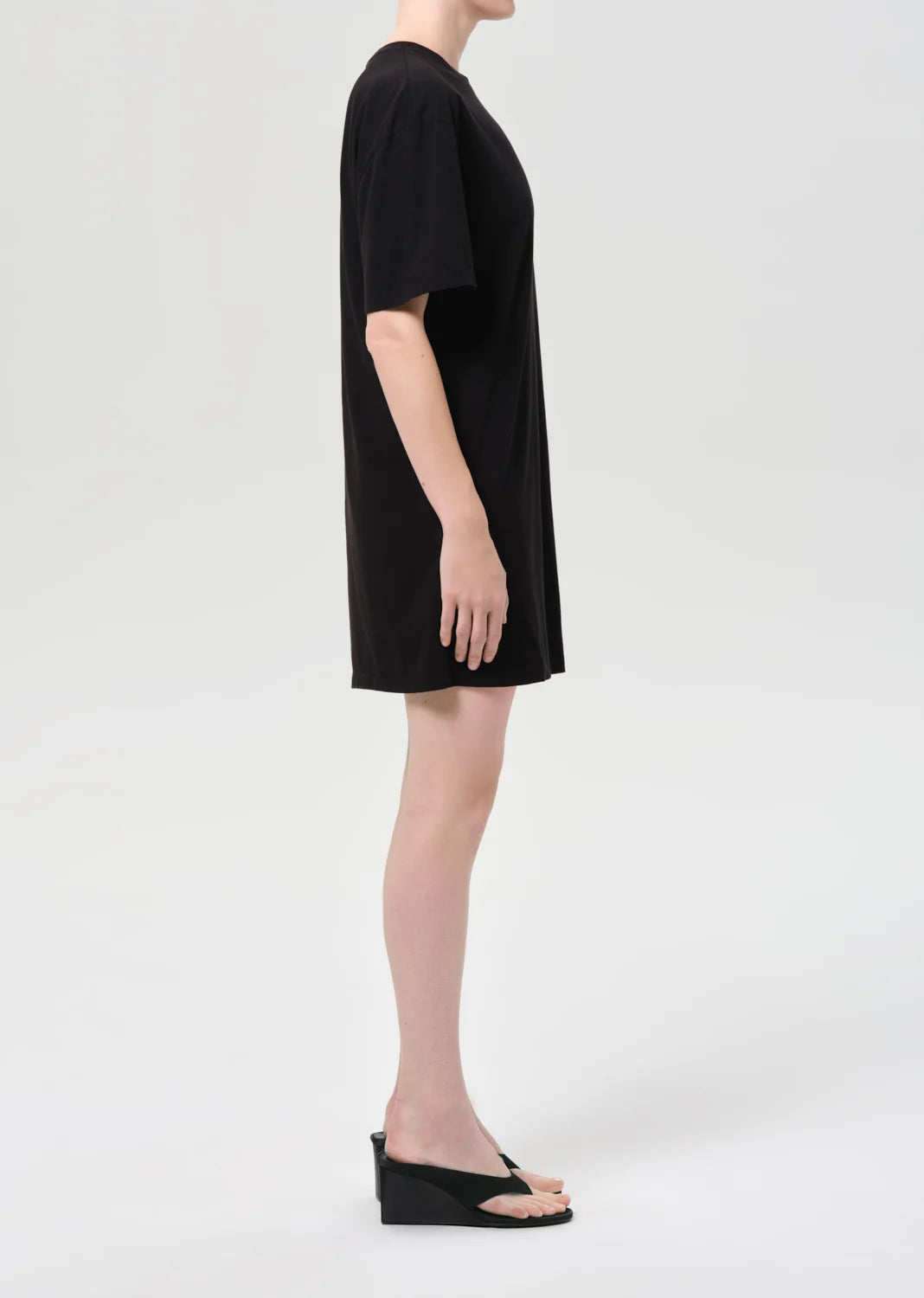 A person stands in profile against a plain background, wearing the Citizens Of Humanity/AGOLDE Roma Dress, a loose black mini dress crafted from organic cotton, paired with black wedge sandals for a simple, casual look.
