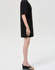 A person stands in profile against a plain background, wearing the Citizens Of Humanity/AGOLDE Roma Dress, a loose black mini dress crafted from organic cotton, paired with black wedge sandals for a simple, casual look.
