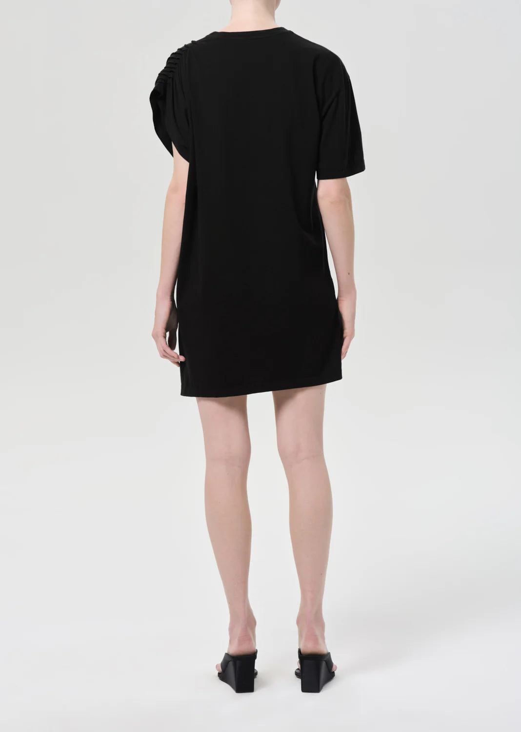 A person faces away from the camera, wearing the Roma Dress by Citizens Of Humanity/AGOLDE— a sleek black mini with short sleeves—paired with heeled black sandals against a plain light background.