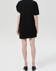 A person faces away from the camera, wearing the Roma Dress by Citizens Of Humanity/AGOLDE— a sleek black mini with short sleeves—paired with heeled black sandals against a plain light background.