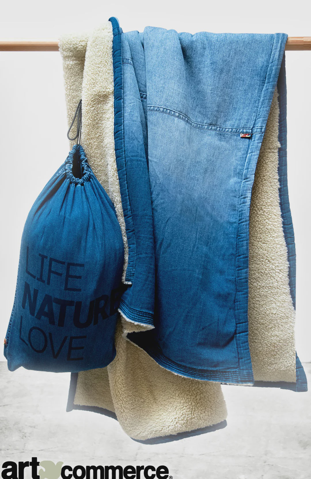 The Free City (sparrow, LLC) Indigo Sherpa Blanket/Indigo Blues, featuring soft white sherpa lining, is draped over a rod beside a blue "LIFE NATURE LOVE" bag. Part of the art commerce logo appears at the bottom.