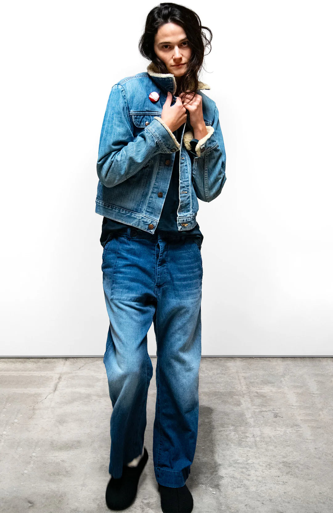 A person stands indoors against a plain white wall, wearing a blue denim jacket with a shearling collar, an Indigo French Sailor Pant by Free City (sparrow, LLC), a black shirt, and black shoes, with hands holding the jacket collar.