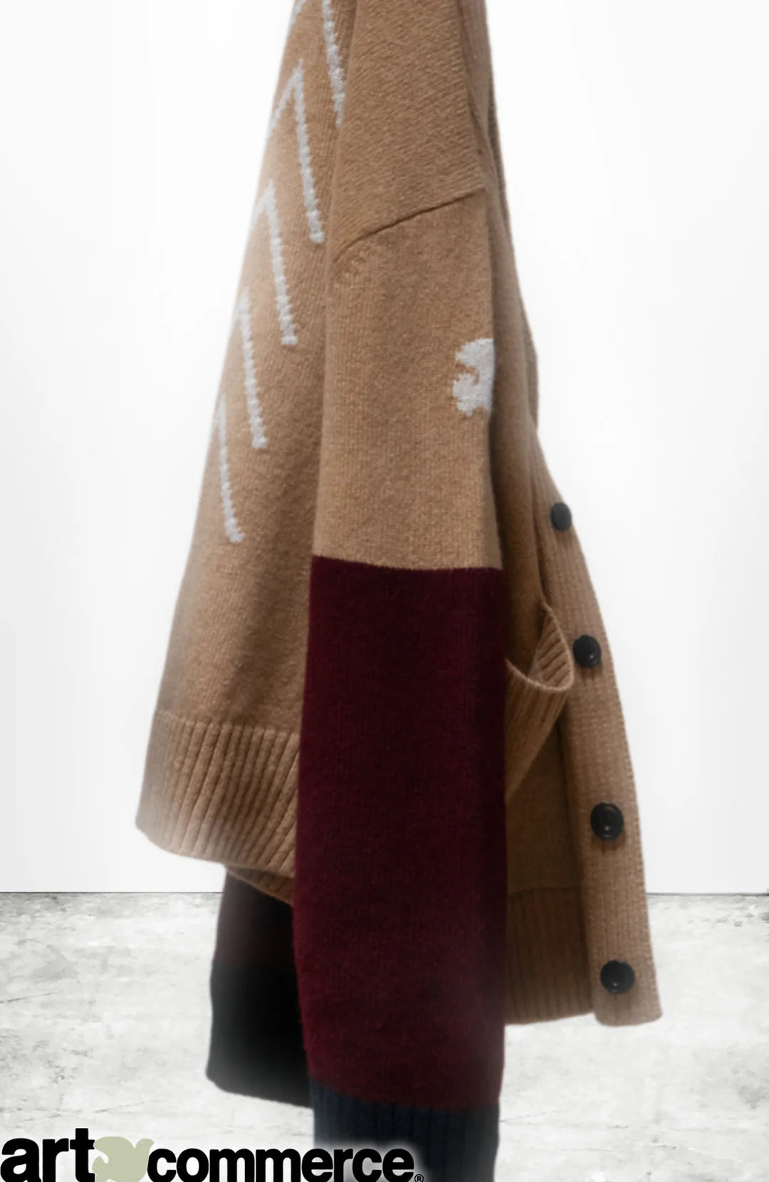 A tan and maroon Cashmere Lux Cardigan by Free City (sparrow, LLC), featuring white patterns, hangs on a hook against a white wall. "Art + commerce" appears below, evoking Loro Piana-inspired unisex elegance.