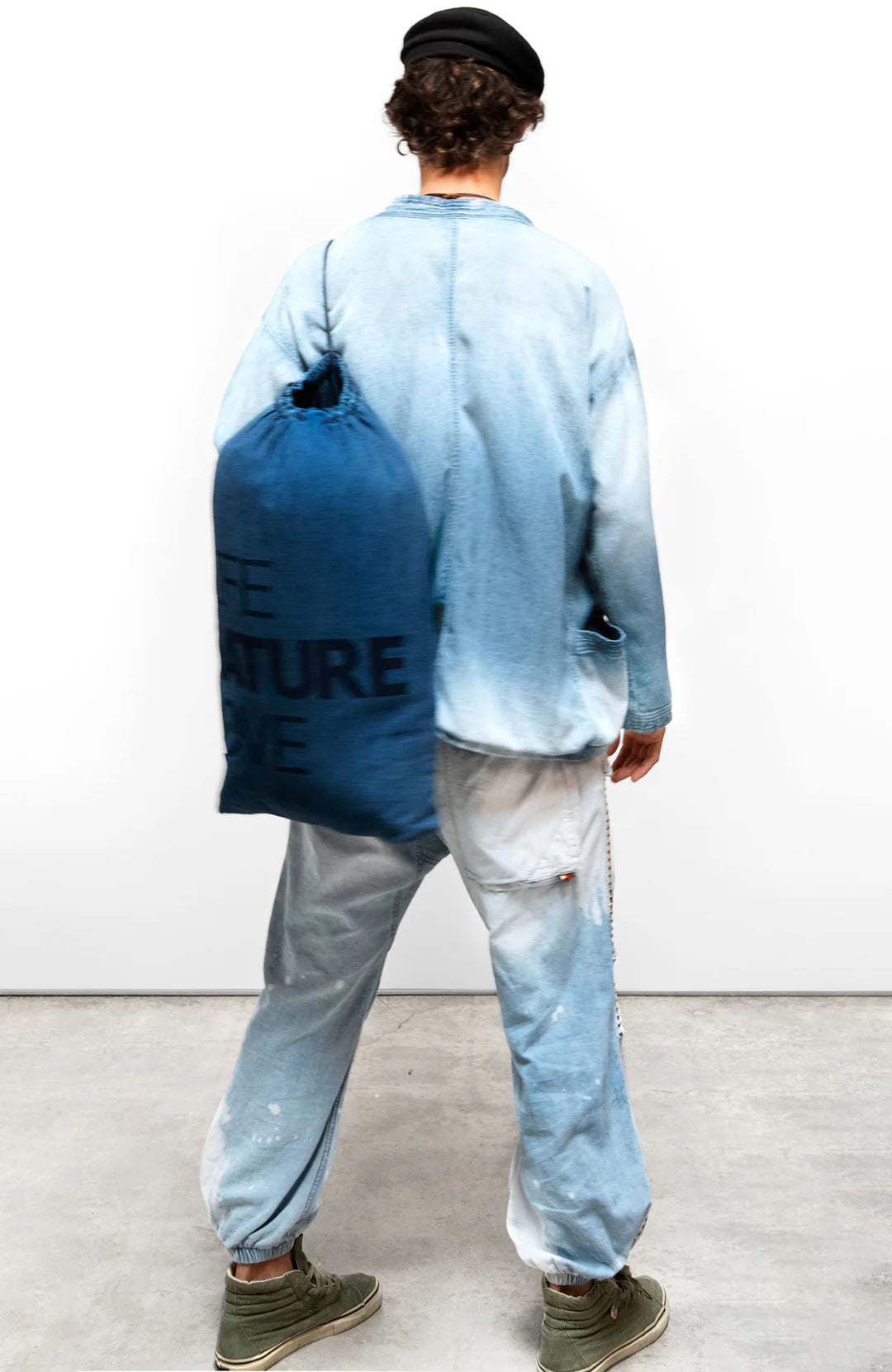 A person in loose indigo clothing and green shoes faces away, carrying a large blue Free City Indigo Sherpa Blanket/Indigo Blues bag with black text and wearing a black beret, set against a plain white background.