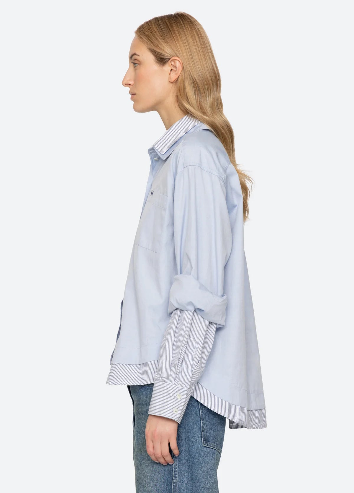 Zayna Button Down shirt in light blue with layered striped hem and cuffs worn with blue jeans, side view.