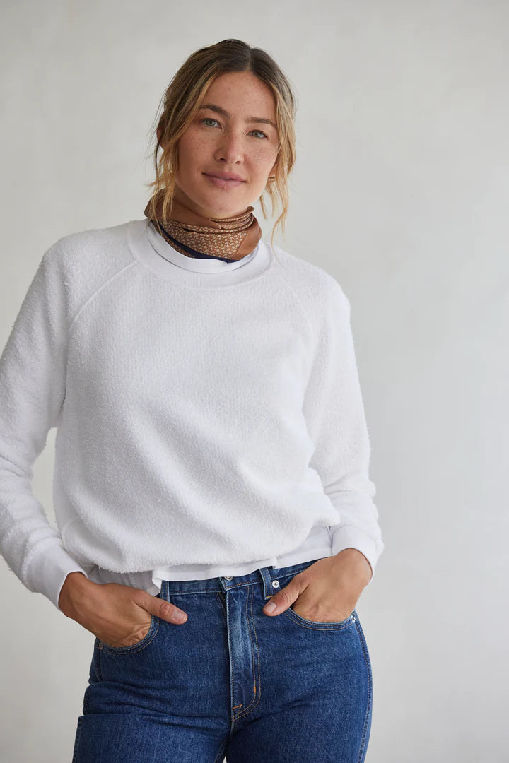 A woman with blonde hair in a loose ponytail wears the Perfectwhitetee Ziggy Reverse LS Sweatshirt and blue jeans, paired with a brown and gold choker necklace, standing against a light gray background with her hands in her pockets.