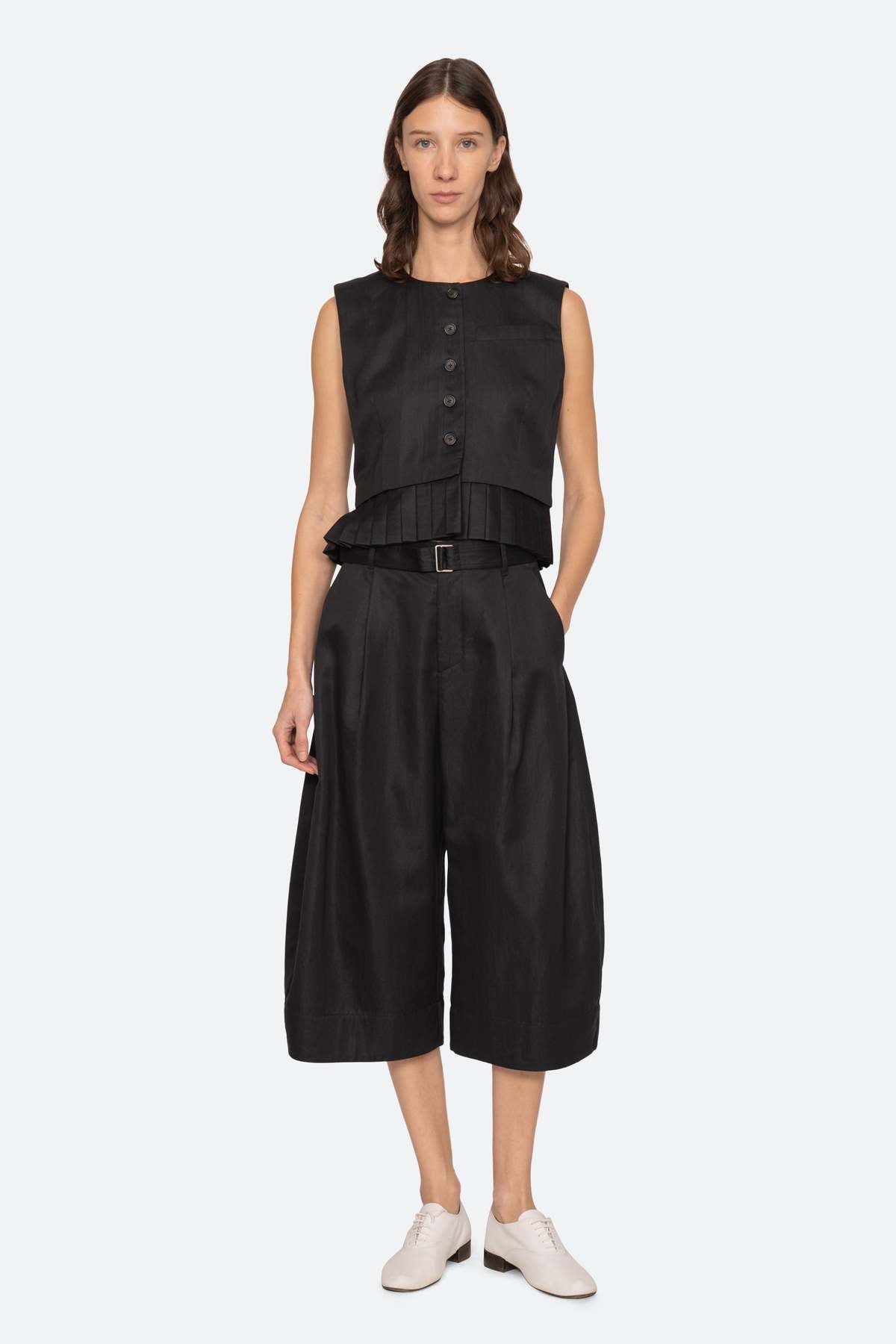 A woman with shoulder-length brown hair and a neutral expression wears the Sea New York Angelina Suiting Culotte, a true-to-size wide-leg black polyester short, styled with a sleeveless peplum top, black belt, and white shoes against a plain background.
