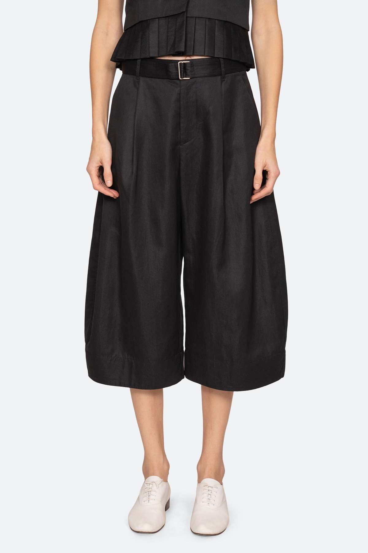 A person stands against a plain light background wearing Sea New York's Angelina Suiting Culotte—loose, wide-leg black polyester pants with a belt—paired with a black top and white shoes.