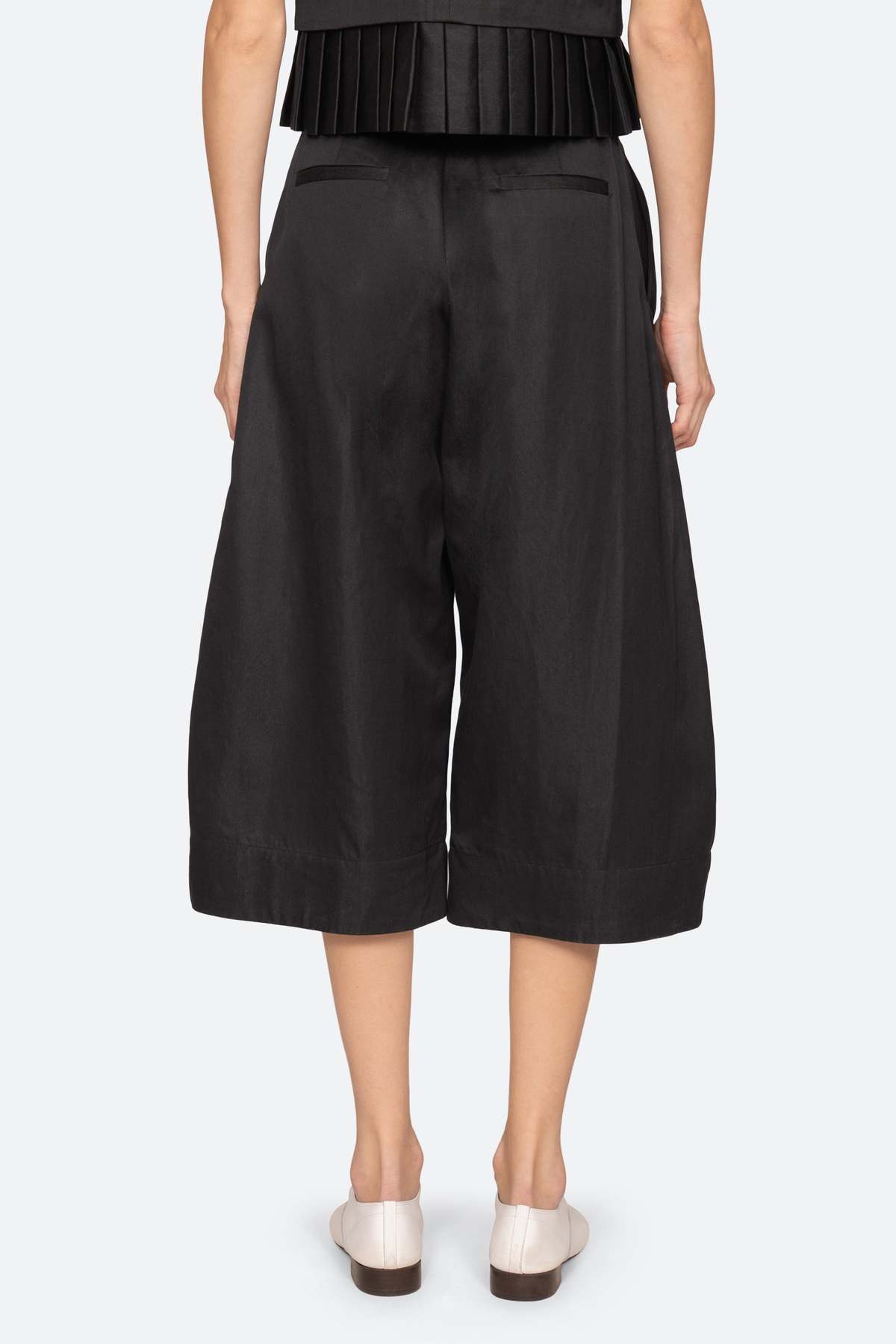 A person is seen from the back wearing Sea New York’s Angelina Suiting Culotte in true-to-size black viscose blend, featuring wide legs, mid-calf crop, back welt pockets, and paired with white shoes against a plain light background.