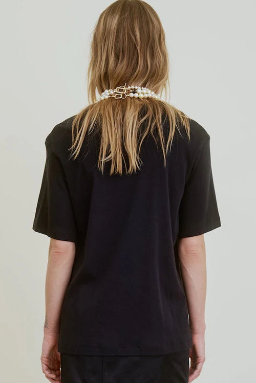 A person with long, light brown hair stands with their back to the camera against a plain light background, wearing the oversized black Campbell OS Crew Tee by Aquarius Cocktail and a layered pearls and gold chain necklace.
