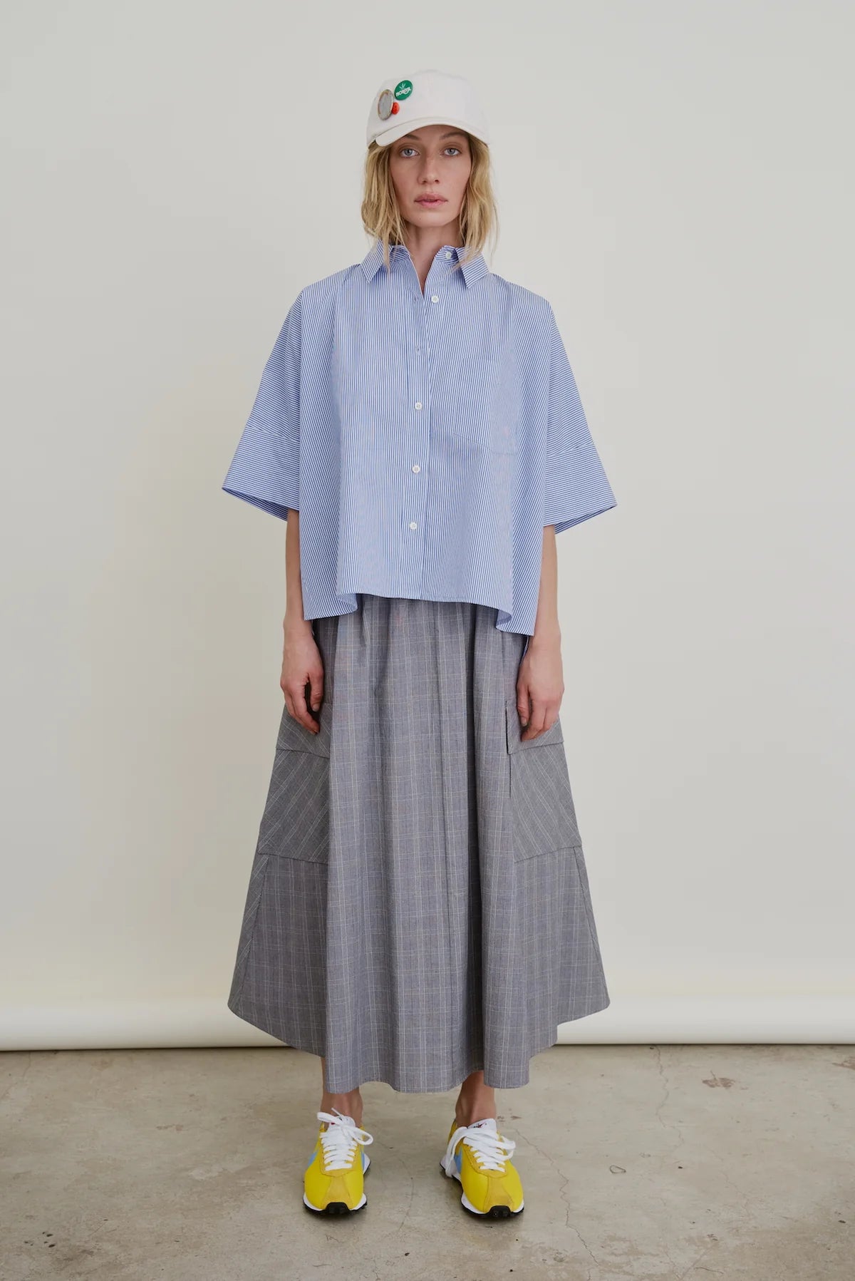 A person stands against a plain background wearing a light blue oversized button-up shirt, yellow sneakers, a white baseball cap with colorful patches, and the Le Cargo Skirt by Aquarius Cocktail—a long gray pleated cargo skirt with an elastic waist.