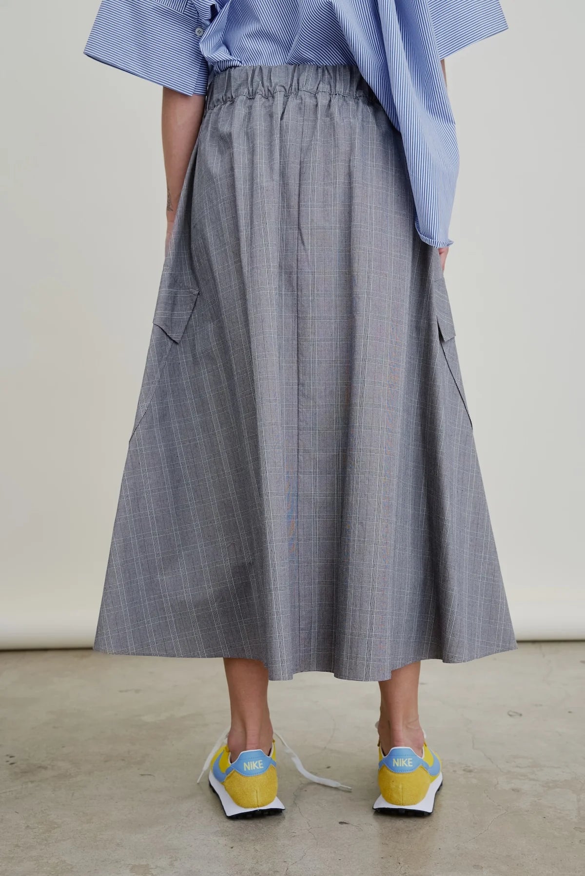 A person stands indoors facing away from the camera, wearing a blue and white striped shirt, yellow Nike sneakers, and the gray Le Cargo Skirt by Aquarius Cocktail with pockets.