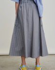 A person stands indoors facing away from the camera, wearing a blue and white striped shirt, yellow Nike sneakers, and the gray Le Cargo Skirt by Aquarius Cocktail with pockets.