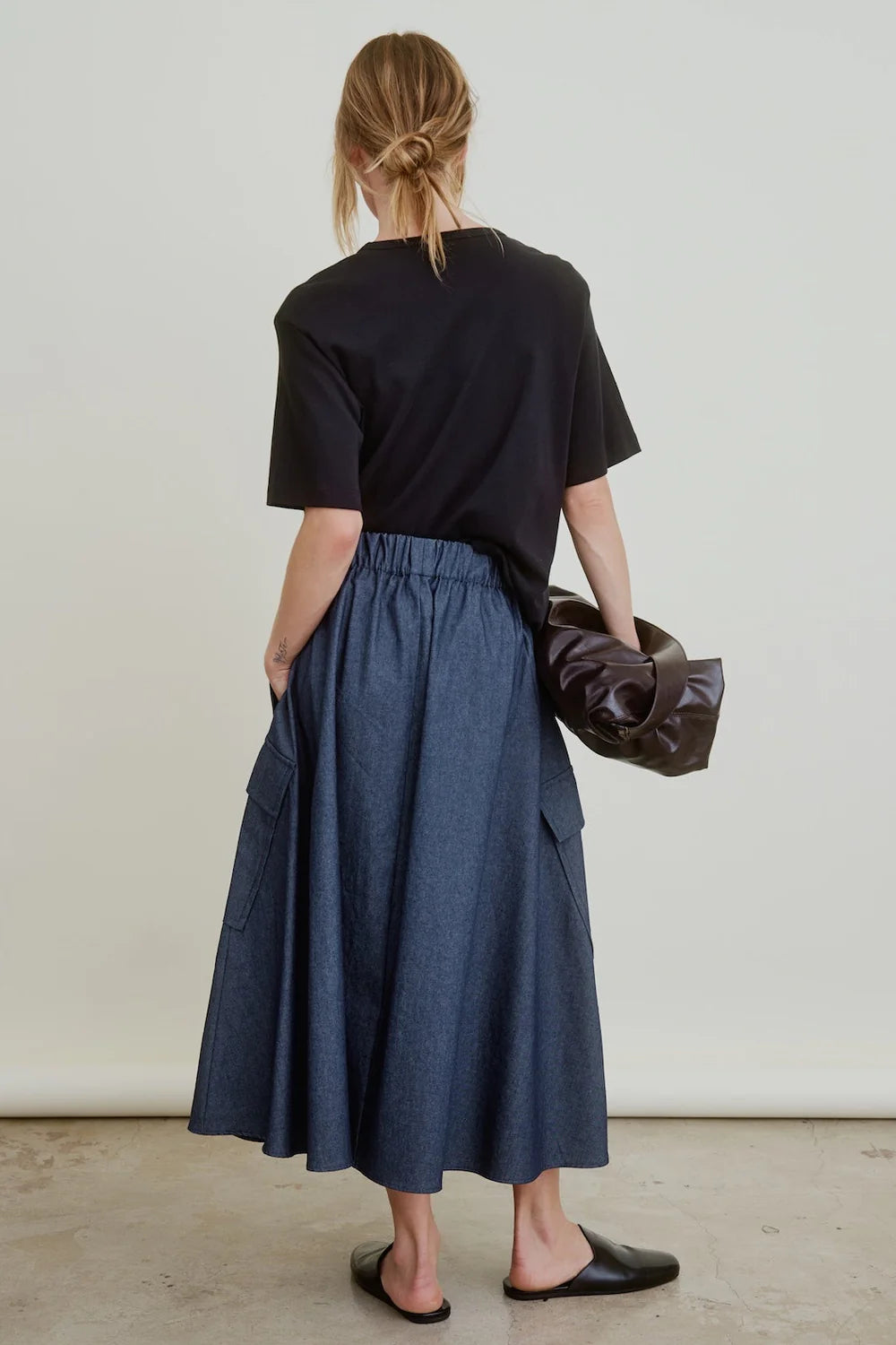 A woman with blonde hair in a low, messy bun wears a black t-shirt, Aquarius Cocktail's Le Cargo Skirt in Japanese cotton poplin denim with oversized pockets and an elastic waistband, black slip-on shoes, and holds a dark brown bag.