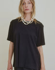 A person with long blonde hair wears the oversized black Campbell OS Crew Tee by Aquarius Cocktail, a black skirt, and layered pearl necklaces, standing against a plain light-colored background.