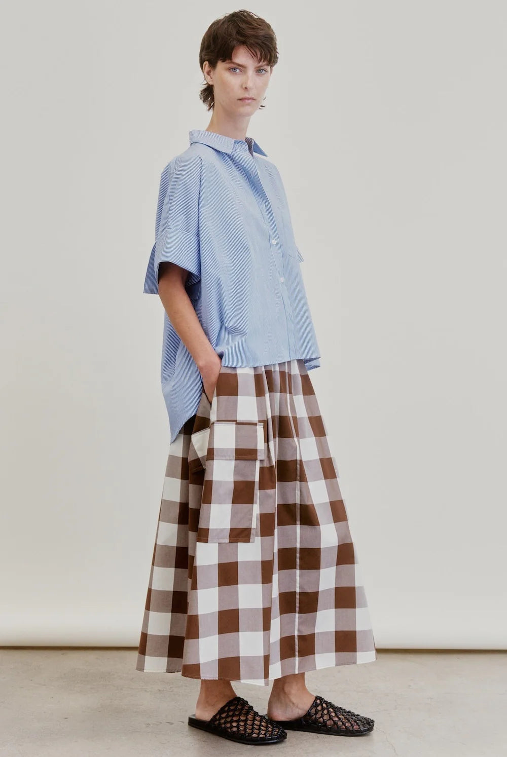 A person stands against a plain background wearing a loose light blue short-sleeve shirt paired with the Aquarius Cocktail Le Cargo Skirt—a long brown and white checkered cargo skirt crafted from Japanese cotton poplin, featuring oversized cargo pockets—and black woven slip-on shoes. Their hands are tucked into the pockets of the skirt.