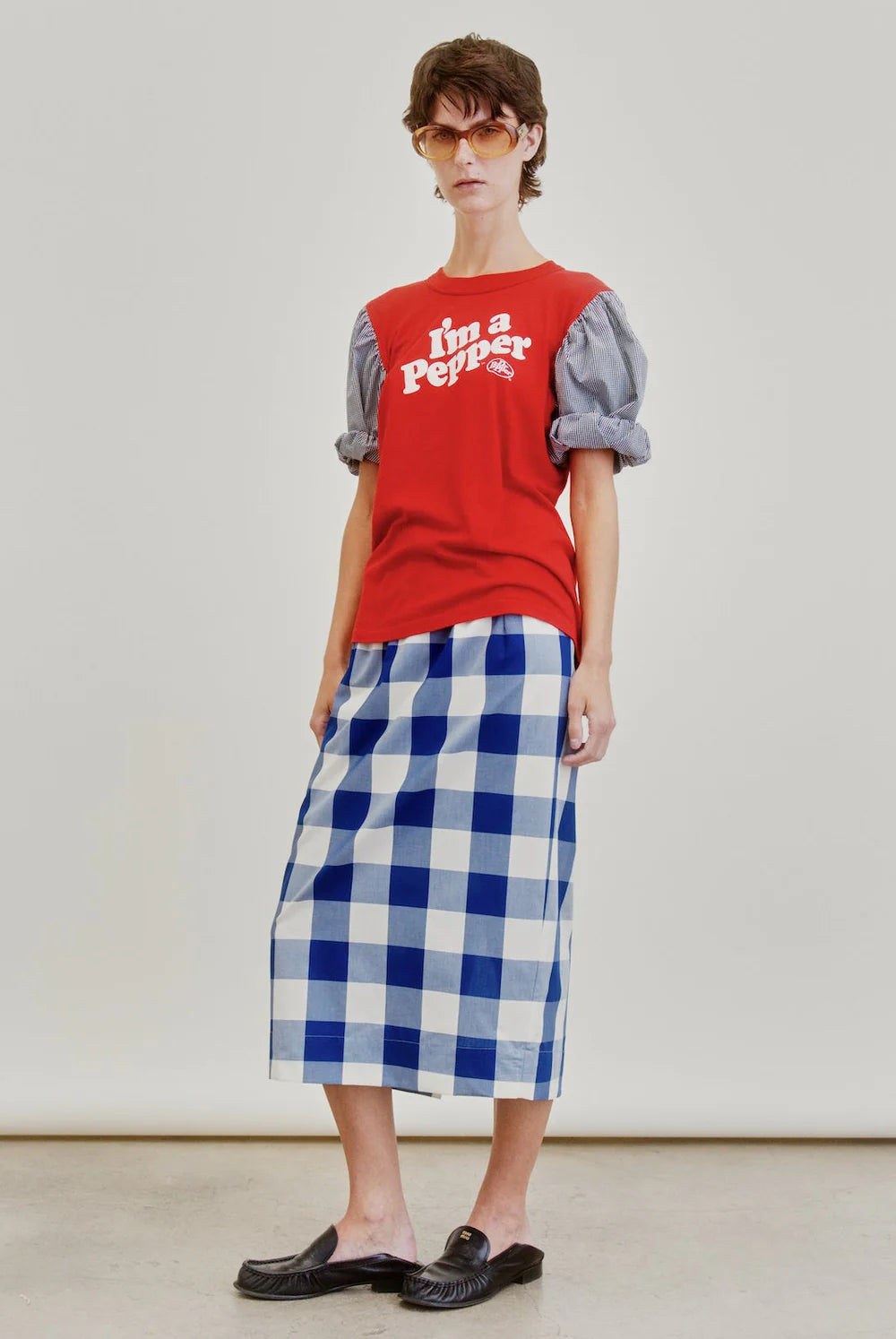 A person wearing a red "I'm a Pepper" T-shirt, a puff-sleeve blouse, an Aquarius Cocktail Le Pencil Slim Skirt in Japanese cotton poplin blue-and-white check, black slip-on shoes, and sunglasses stands against a plain light background.