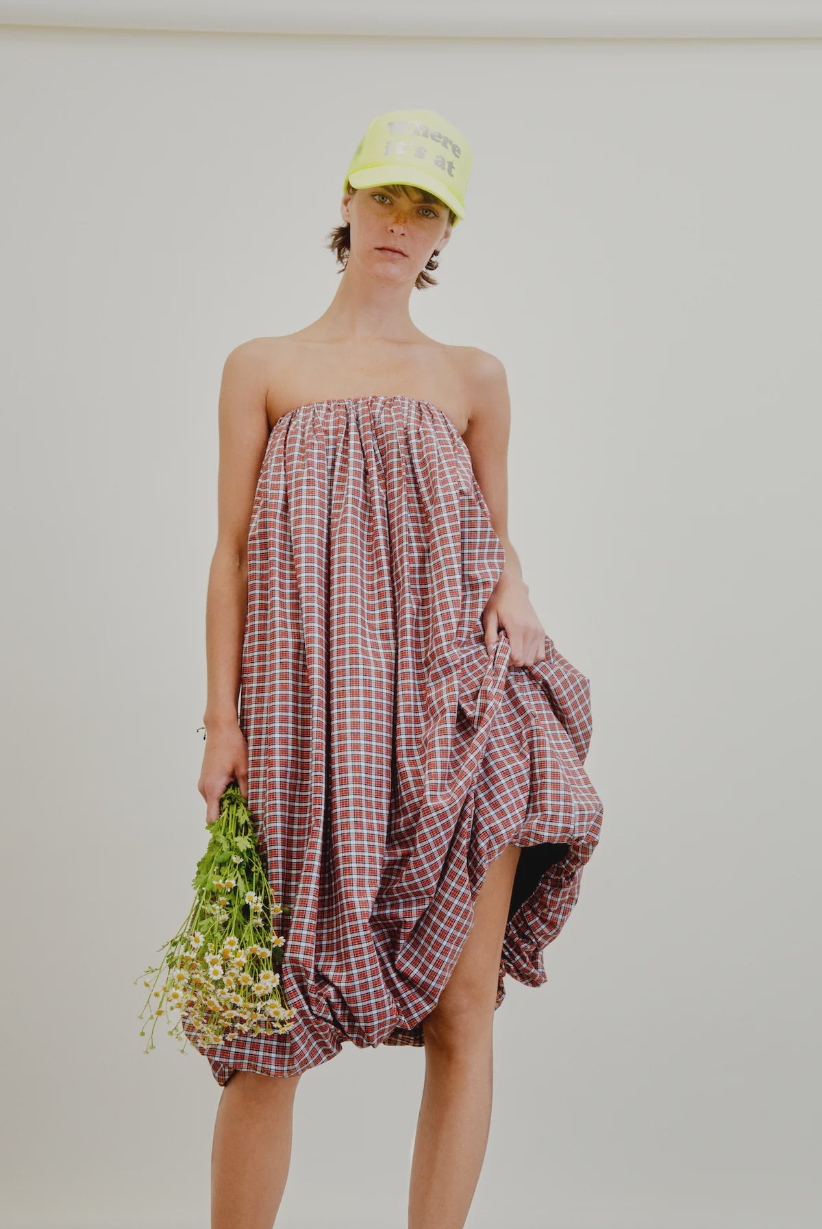 A person models the Aquarius Cocktail Cosmo Bubble Dress—a loose, strapless red and white checkered Japanese cotton piece—paired with a yellow cap and holding small white flowers against a plain white background.