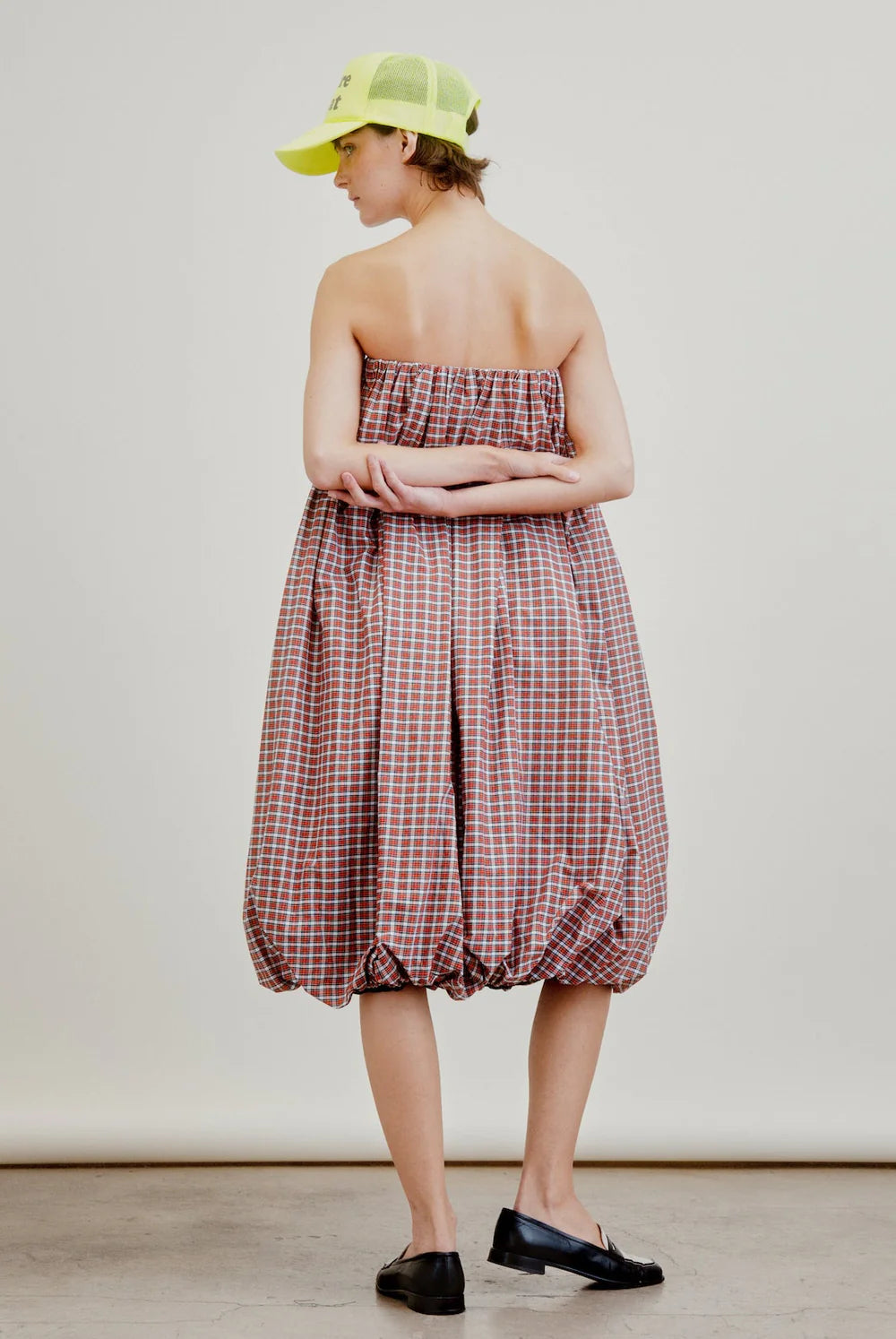A person stands with arms crossed, wearing the red and white checked Cosmo Bubble Dress by Aquarius Cocktail, paired with black loafers and a neon yellow baseball cap against a plain, neutral background.