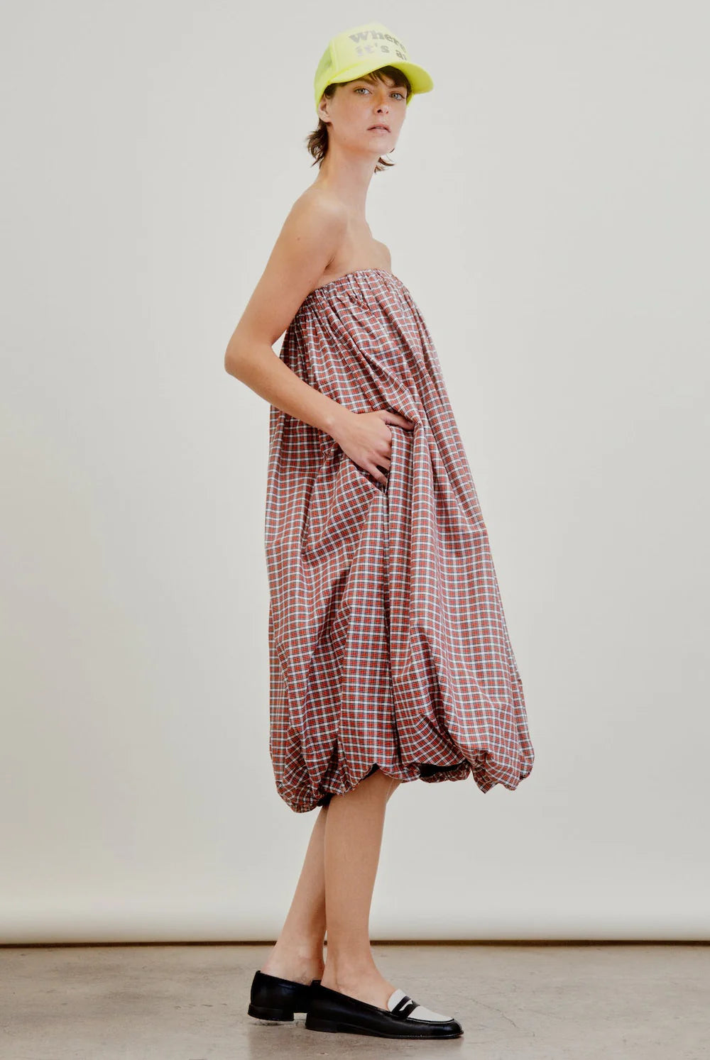 A person wearing the Cosmo Bubble Dress by Aquarius Cocktail, a loose strapless red and white plaid piece, with black loafers and a neon yellow cap, stands in a neutral studio setting.