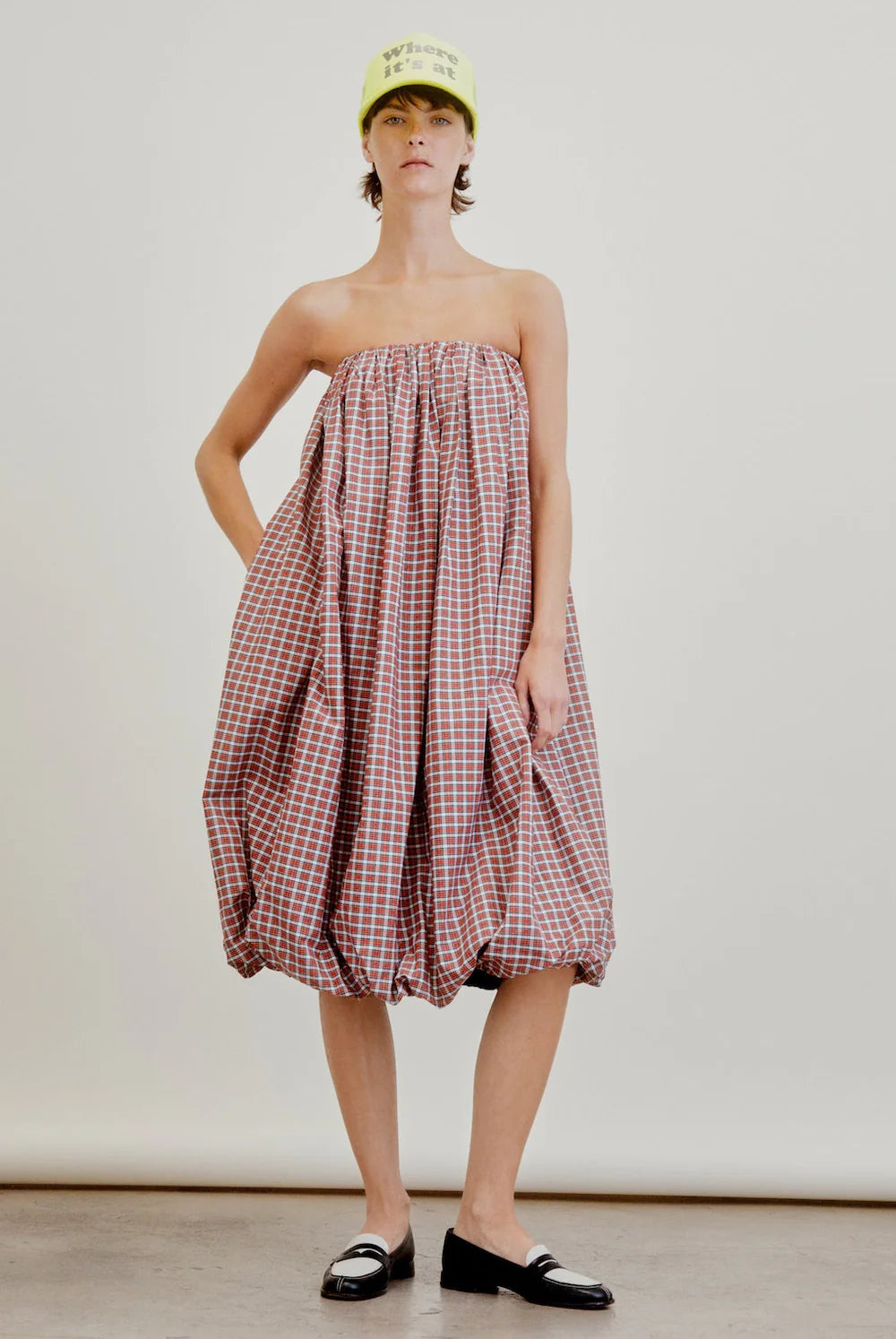 A person wears the Aquarius Cocktail Cosmo Bubble Dress—a loose, strapless red plaid Japanese cotton dress with a gathered hem—paired with black loafers and a neon yellow baseball cap against a plain background.