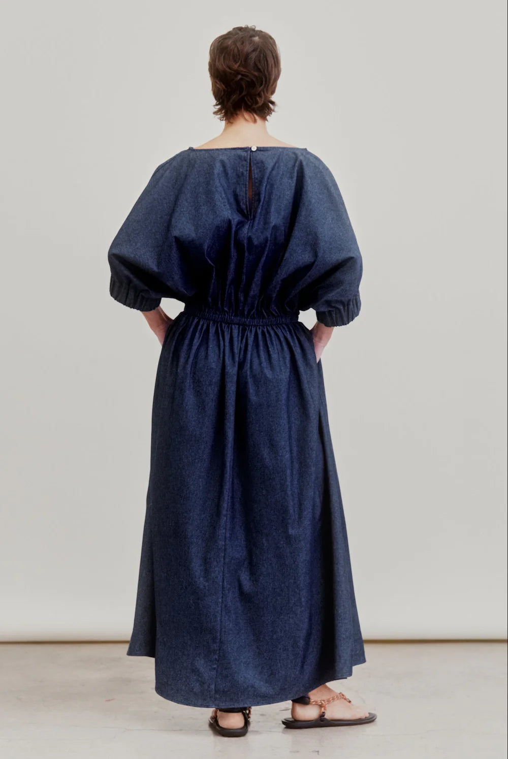 A person faces away, wearing the Aquarius Cocktail French 75 denim dress—loose-fitting, ankle-length with gathered sleeves, waist, and a boat neckline. They have short hair and flat sandals, set against a plain light background.